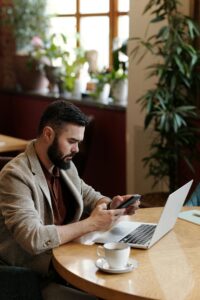 Bearded man using smartphone and laptop at café with coffee, focused on remote work.